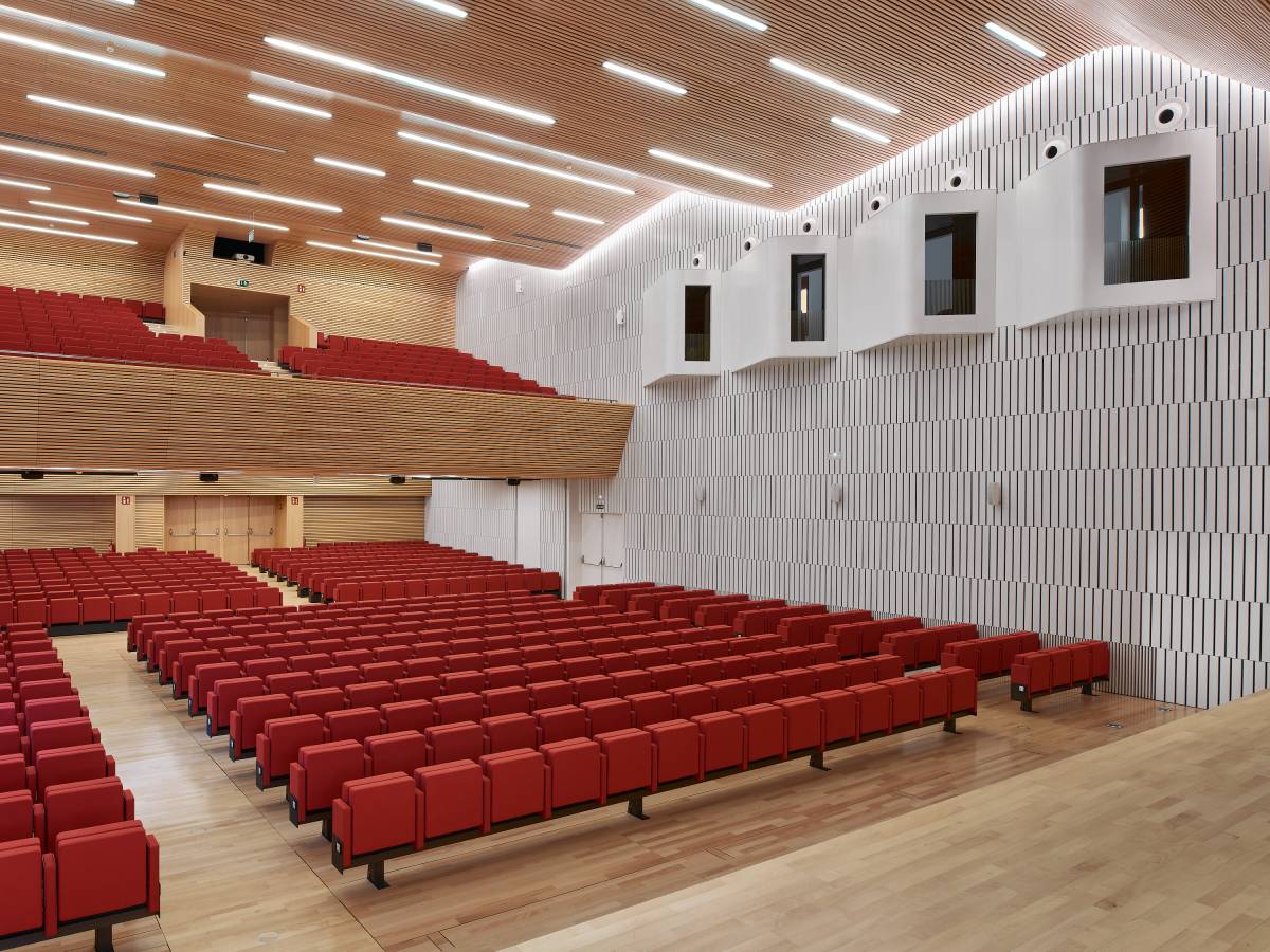 The image shows the interior of a large auditorium in a modern building. The seating is arranged in various sections and floors, with rows of chairs facing the stage. The auditorium is currently empty, but is designed to accommodate a large number of people, making it suitable for a variety of events and conferences. In the background, the white wall shows 4 simultaneous translation booths facing the stage. The architecture and interior design are impressive, highlighting the attention to detail and the use of natural light, which creates a welcoming and comfortable atmosphere.