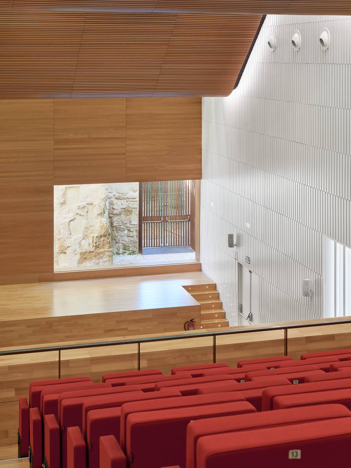 Interior view of the Congress Centre of Cordoba, Spain, designed by LAP architects, featuring a wooden ceiling, a stage with red seats and a view of a stone wall through a window.