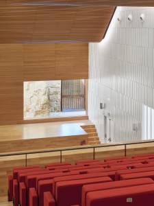Interior view of the Congress Centre of Cordoba, Spain, designed by LAP architects, featuring a wooden ceiling, a stage with red seats and a view of a stone wall through a window.