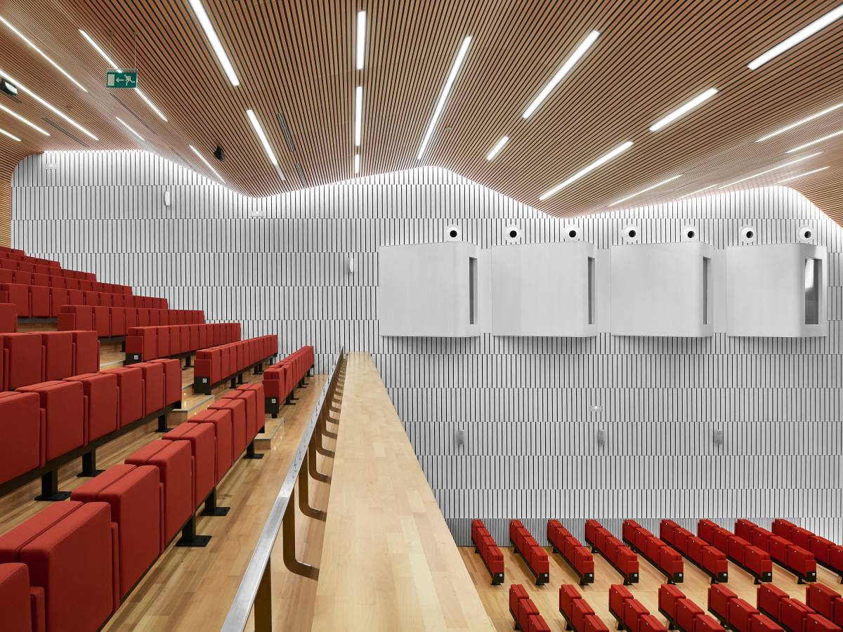 The image shows the interior of a large auditorium in a modern building. The seating is arranged in various sections and floors, with rows of chairs facing the stage. The auditorium is currently empty, but is designed to accommodate a large number of people, making it suitable for a variety of events and conferences. In the background, the white wall shows 4 simultaneous translation booths facing the stage. The architecture and interior design are impressive, highlighting the attention to detail and the use of natural light, which creates a welcoming and comfortable atmosphere.